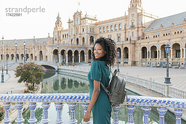 Lächelnde junge Frau mit lockigem Haar steht am Geländer an der Plaza De Espana  Sevilla  Spanien