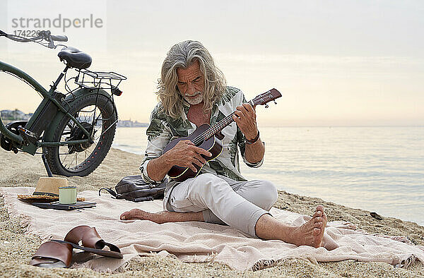 Reifer Mann mit grauen Haaren spielt am Strand Ukulele