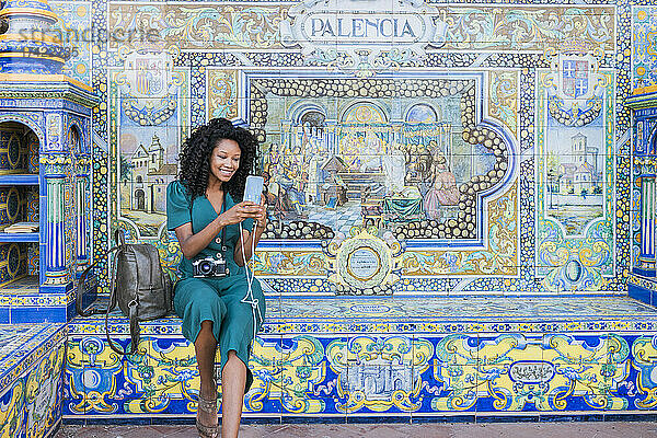 Lächelnde junge Frau  die ihr Smartphone benutzt  während sie vor einem Mosaik auf der Plaza De Espana in Sevilla  Spanien  sitzt