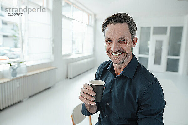 Lächelnder Geschäftsmann  der am Arbeitsplatz Kaffee im Einwegbecher trinkt
