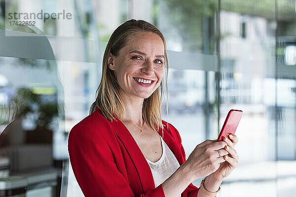 Lächelnde Geschäftsfrau hält Mobiltelefon vor Glaswand