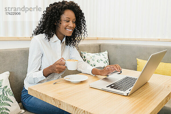 Lächelnde Geschäftsfrau trinkt Kaffee  während sie im Café an einem Videoanruf über ihren Laptop teilnimmt