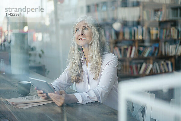 Nachdenkliche Geschäftsfrau mit digitalem Tablet  die durch das Caféfenster schaut
