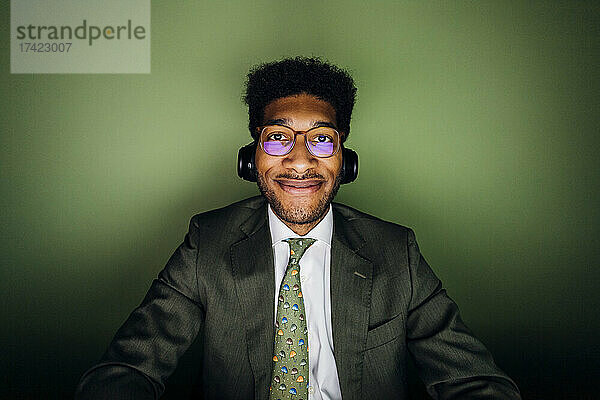 Lächelnder Geschäftsmann mit Brille und kabellosen Kopfhörern vor grüner Wand