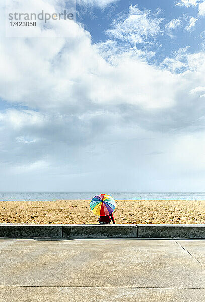 Frau sitzt an sonnigen Tagen mit buntem Sonnenschirm am Strand