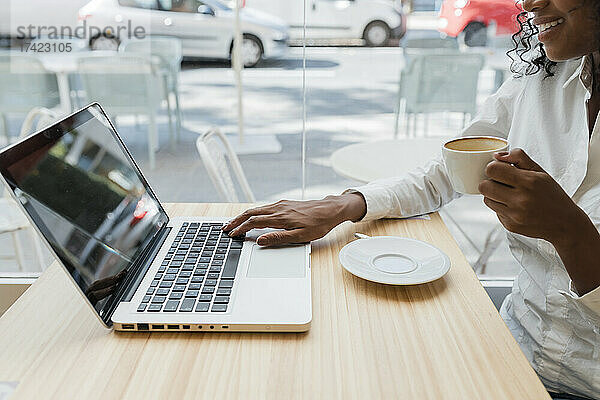 Freiberuflerin hält Kaffeetasse in der Hand  während sie Laptop im Café benutzt