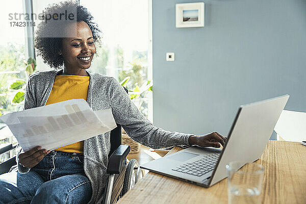 Junge Freiberuflerin hält ein Dokument in der Hand  während sie im Heimbüro am Laptop arbeitet