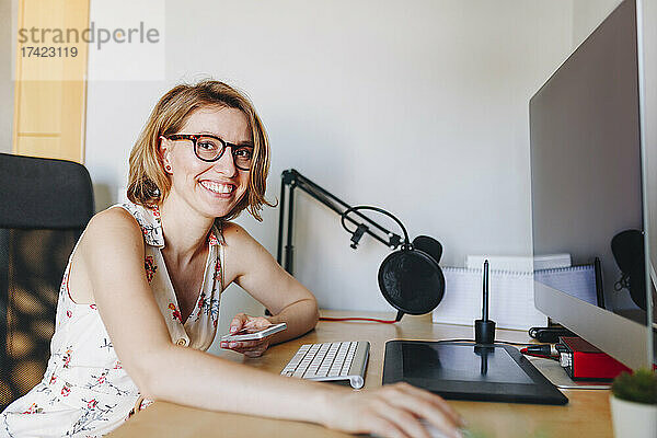 Lächelnde Geschäftsfrau hält Mobiltelefon in der Hand  während sie im Heimbüro am Computertisch sitzt