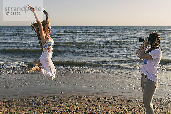 Frau fotografiert Freundin beim Springen am Strand