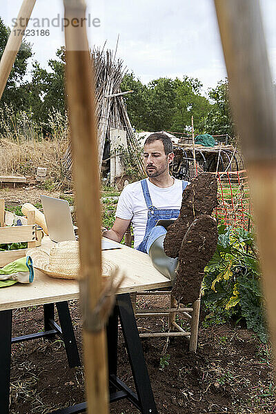 Landwirt benutzt Laptop  während er mit erhobenen Füßen am Tisch im landwirtschaftlichen Feld sitzt