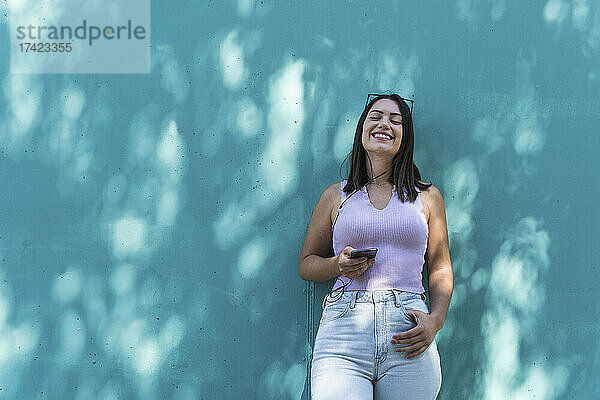 Lächelnde junge Frau mit Mobiltelefon lehnt an der Wand