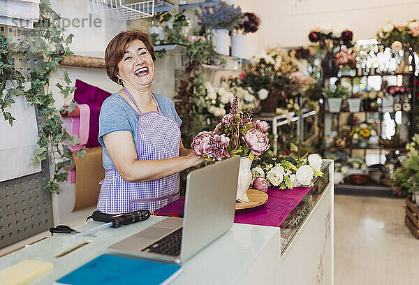 Fröhliche Unternehmerin mit braunen Haaren arrangiert Blumen  während sie im Geschäft steht