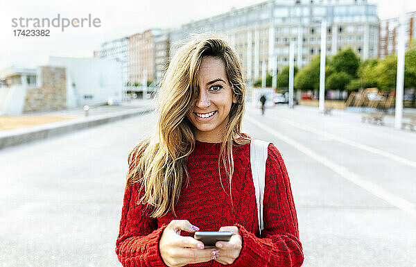 Lächelnde Frau hält Smartphone in der Hand  während sie auf der Straße steht