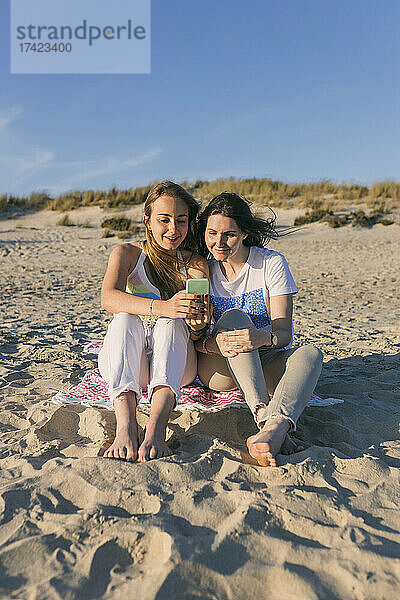 Junge Freundinnen nutzen ihr Smartphone  während sie am Strand sitzen