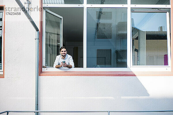 Lächelnder junger Geschäftsmann hält Kaffeetasse in der Hand  während er am Fenster steht