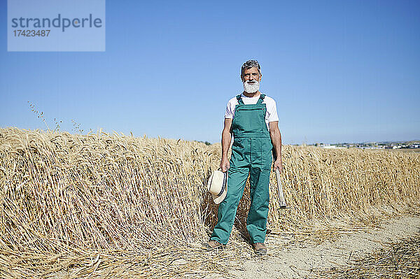 Männlicher Landarbeiter steht an sonnigen Tagen mit Hut und Hacke auf dem Weizenfeld