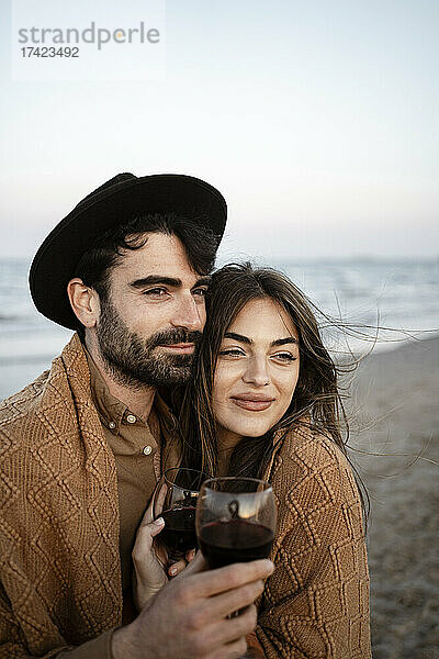 Lächelndes Paar in Decke gehüllt und mit Weinglas am Strand