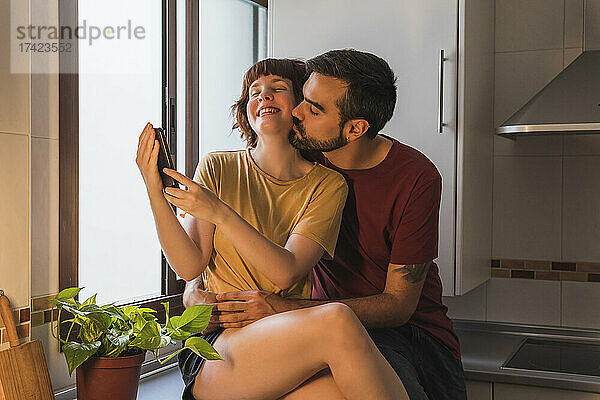 Lächelnde Frau macht ein Selfie mit ihrem Freund  der sich zu Hause in der Küche küsst