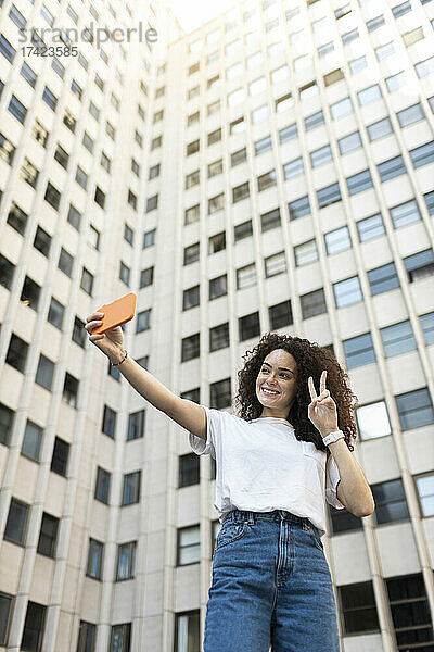 Junge Frau macht ein Friedenszeichen  während sie vor dem Gebäude ein Selfie macht