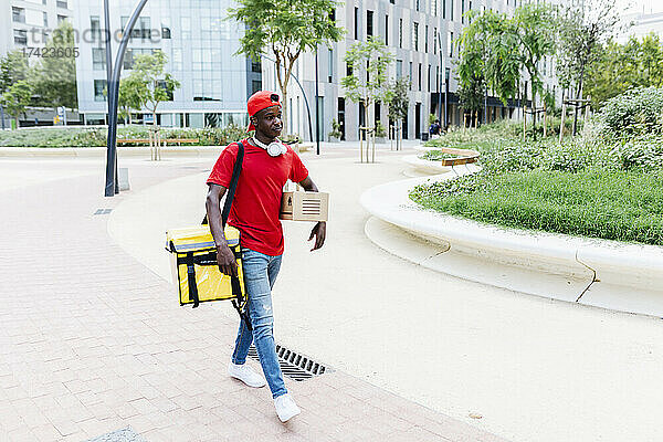Junger Zusteller mit Rucksack und Paket läuft auf Fußweg in der Stadt