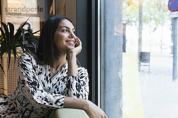 Lächelnde junge Frau träumt  während sie durch das Fenster im Café schaut