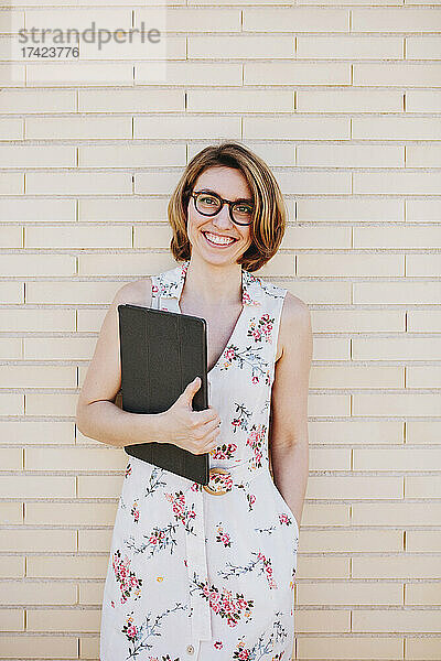 Lächelnde Geschäftsfrau mit Brille und digitalem Tablet steht vor einer Ziegelwand