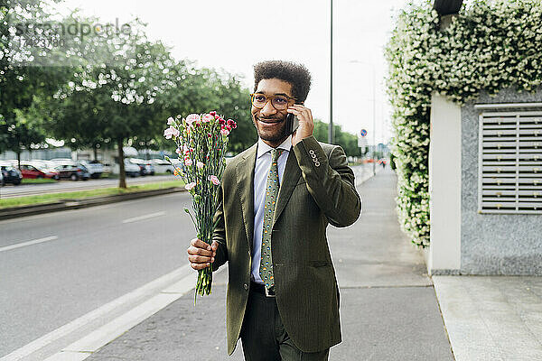 Lächelnder Geschäftsmann hält einen Blumenstrauß in der Hand  während er auf der Straße mit dem Handy spricht