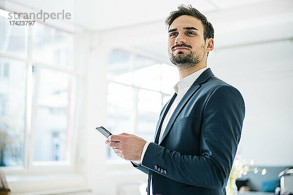 Junger männlicher Berufstätiger schaut weg  während er sein Smartphone im Büro hält