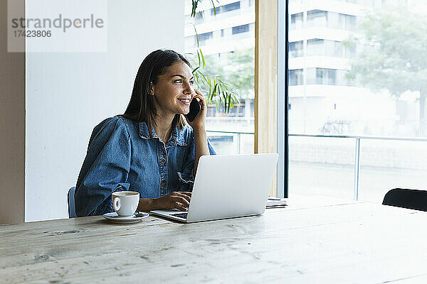 Lächelnde Geschäftsfrau telefoniert  während sie mit einem Laptop im Café sitzt