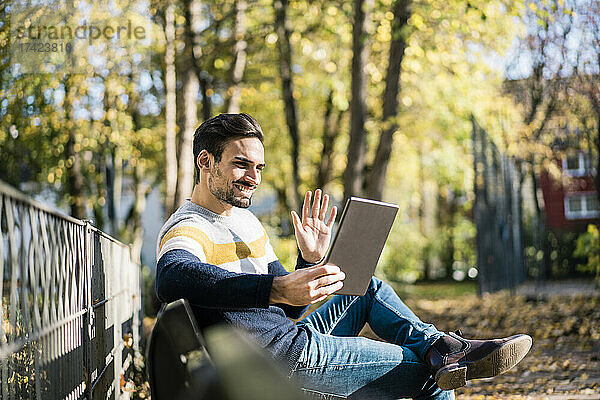 Junger Mann winkt mit der Hand  während er an einem sonnigen Tag im Park einen Videoanruf über ein digitales Tablet tätigt