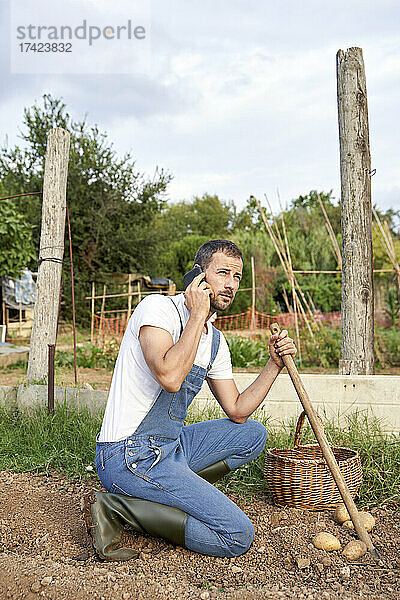 Landarbeiter hält Gartenhacke in der Hand und schaut weg  während er auf dem landwirtschaftlichen Feld mit dem Smartphone spricht