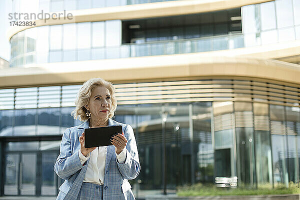Nachdenkliche Geschäftsfrau hält digitales Tablet in der Hand  während sie vor dem Bürogebäude steht