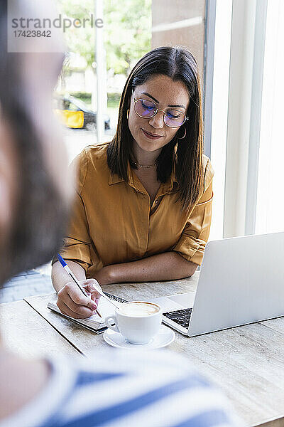 Junge berufstätige Frau schreibt Tagebuch  während sie mit einem männlichen Kollegen im Café sitzt