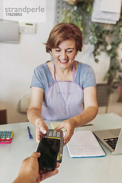 Lächelnde reife Floristin hält Kreditkartenlesegerät in der Hand  während der Kunde an der Kasse kontaktlos per Mobiltelefon bezahlt