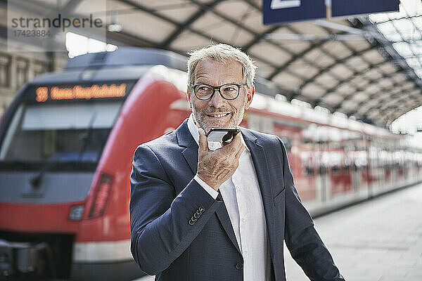 Lächelnder männlicher Berufstätiger  der wegschaut  während er am Bahnhof Voicemail über sein Smartphone sendet