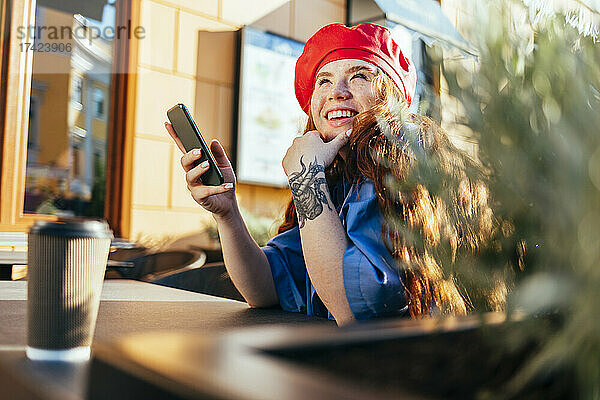Glückliche Frau mit Smartphone  die im Straßencafé nachdenkt