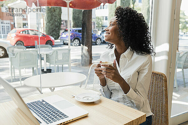 Lächelnde Geschäftsfrau hält Kaffeetasse in der Hand  während sie durch das Caféfenster schaut