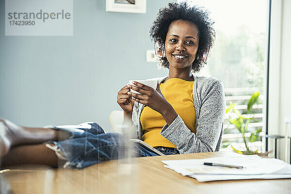 Lächelnde Frau hört Musik  während sie am Tisch im Wohnzimmer Kaffee trinkt