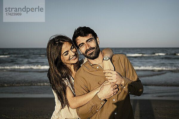 Lächelnde Frau umarmt Mann von hinten am Strand