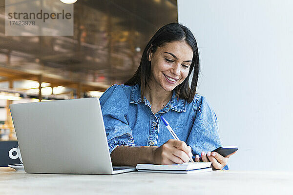 Lächelnde Geschäftsfrau  die im Café Tagebuch schreibt  während sie ihr Mobiltelefon in der Hand hält
