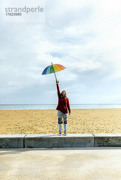 Frau hält Regenschirm mit erhobener Hand  während sie vor bewölktem Himmel am Strand steht