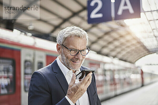 Lächelnder Geschäftsmann  der am Bahnsteig Voicemail per Mobiltelefon sendet