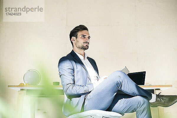 Nachdenklicher junger männlicher Berufstätiger sitzt mit digitalem Tablet auf einem Stuhl im Büro