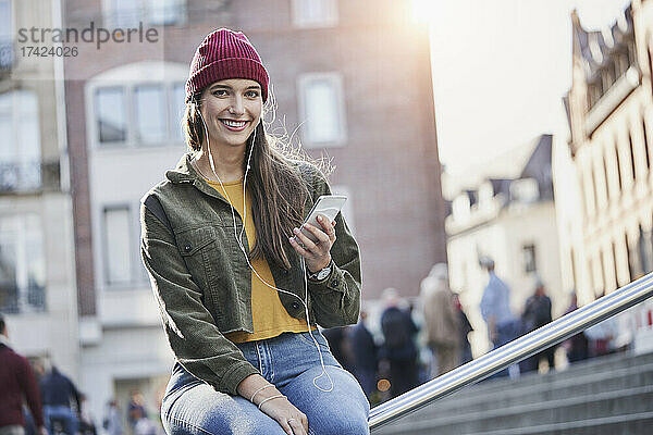 Schöne junge Frau hört Musik  während sie auf dem Geländer sitzt