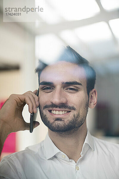 Lächelnder junger Geschäftsmann  der durch Glas auf einem Smartphone spricht