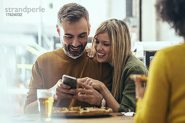 Glückliche männliche und weibliche Freunde  die im Restaurant ihr Smartphone teilen