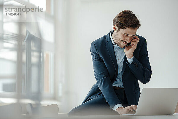 Junger Geschäftsmann benutzt Laptop  während er im Büro mit dem Mobiltelefon telefoniert