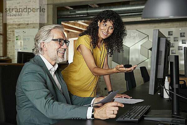 Lächelnde Geschäftsfrau gestikuliert  während sie am Computer mit einem männlichen Kollegen im Büro diskutiert