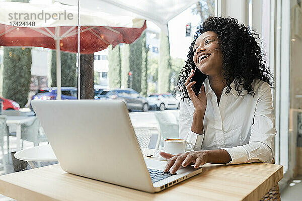 Glückliche Geschäftsfrau mit Laptop  die im Café auf dem Smartphone spricht