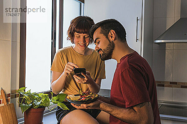 Lächelnde Frau fotografiert Essen  das ihr Freund zu Hause in der Küche hält